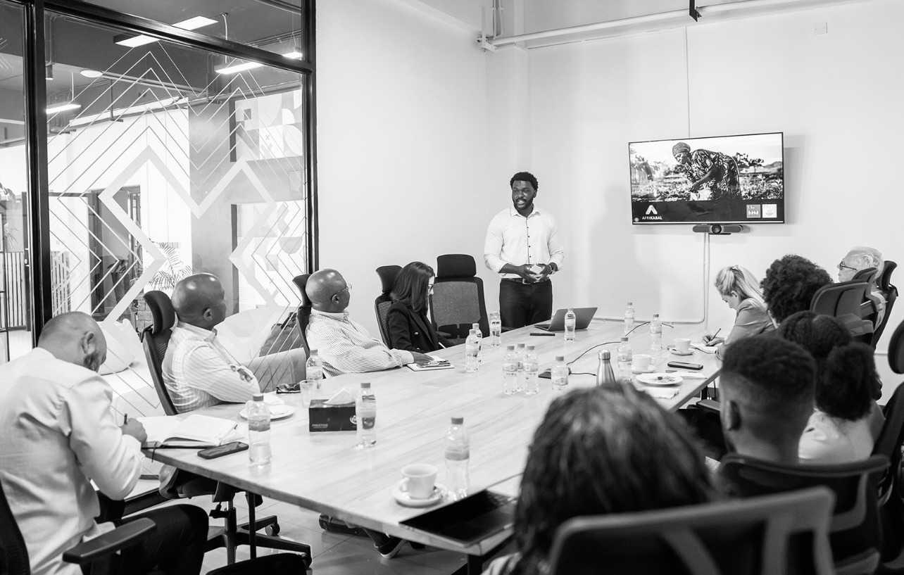 Tejiri Jesse standing at the head of a boardroom table, presenting AXK Network to a room of partners.